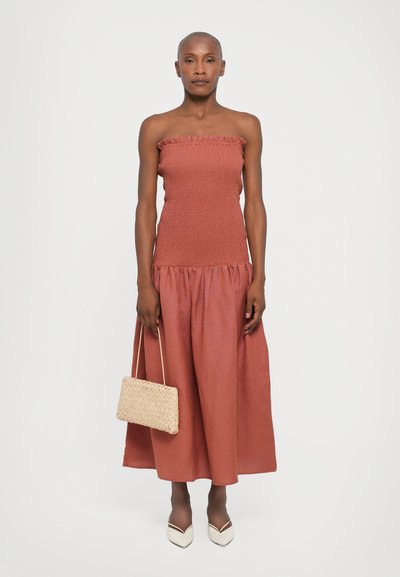 Strapless, rust-colored dress with gathered bodice and flowing skirt. Woven beige handbag and white pointed-toe shoes complete the look.