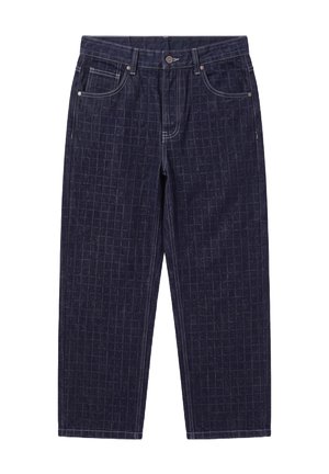 Homme aux cheveux courts et à la barbe, portant une veste en jean sombre à motifs et un pantalon avec des baskets blanches, debout les mains dans les poches.