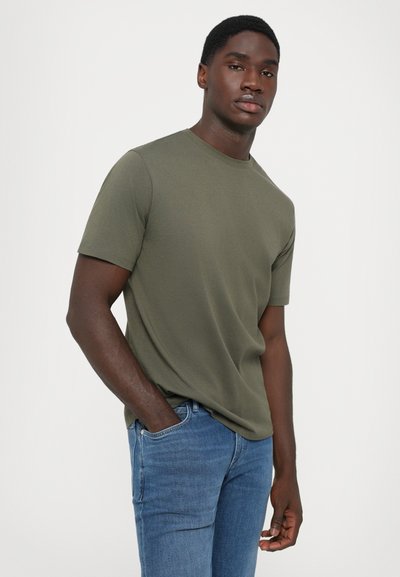 Tânăr îmbrăcat cu un tricou cu m�ânecă scurtă de culoare verde măsliniu și blugi, stând cu o mână în buzunar, pe un fundal deschis simplu.