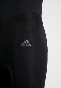 Leggings de sport noirs en tissu extensible, avec une texture lisse. Comprend un petit logo Adidas en argent sur le côté.