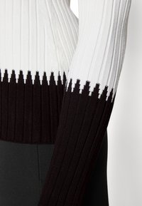 Haut à manches longues en maille côtelée avec sections supérieures blanches et inférieures noires séparées par un motif à bords dentelés autour de la taille et du bras.