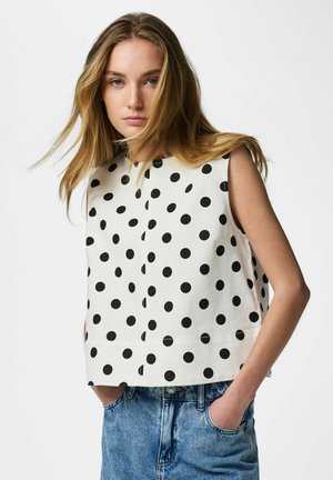 Młoda kobieta z długimi blond włosami, ubrana w bezrękawnik w białym kolorze w czarne kropki i niebieskie dżinsy, ręce w kieszeniach, neutralne tło.