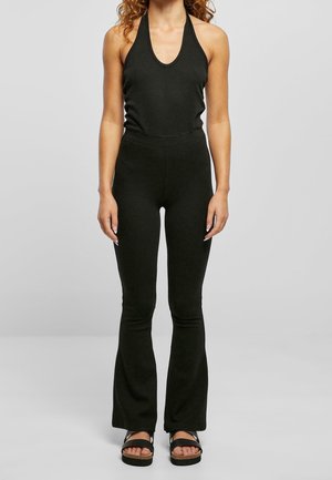 Kobieta ubrana w czarną bluzkę na szyję typu halter, dopasowane spodnie z wysokim stanem i rozszerzanymi nogawkami oraz czarne sandały na platformie, stojąca na jednolitym tle.