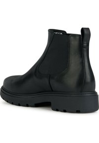 Bota de tobillo de cuero negro con paneles laterales elásticos, puntera redonda y suela de goma texturizada. Cuenta con una lengüeta para facilitar su uso.