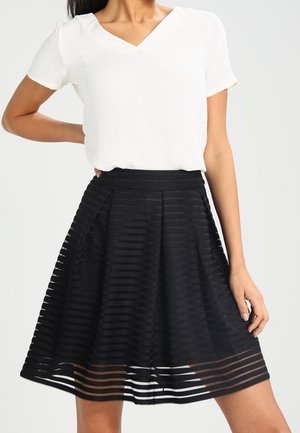 Vrouw draagt een wit kortärmelig V-hals shirt en een zwarte plissé rok tot op de knie met horizontale doorschijnende strepen.