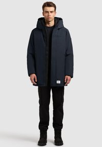 Mørkeblå vandtæt parka-jakke med hætte, frontlynlås og to sidelommer. Båret over en sort skjorte og parret med sorte cargo-bukser.