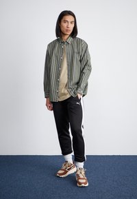 Camisa de botón a rayas verdes sobre una camiseta beige, pantalones negros con rayas blancas y zapatillas marrones con detalles blancos.
