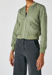 Veste bomber courte vert olive avec fermeture éclair frontale et poches à rabat, portée avec un jean noir délavé taille haute sur une personne debout.