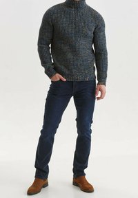 Pull à col roulé en maille aux tons bleus et dorés, présentant un motif texturé, associé à un jean foncé et des bottines marron.