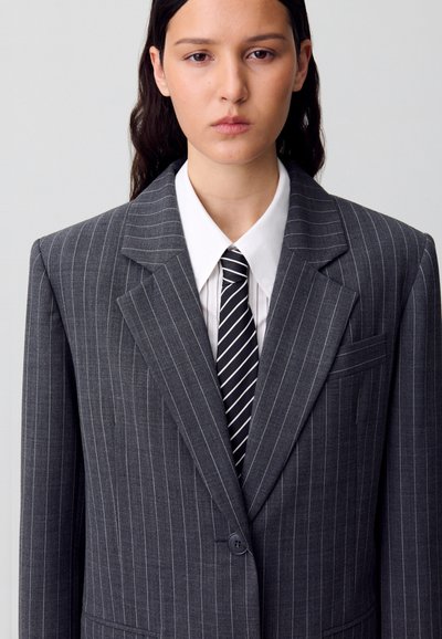 Blazer gris a rayas con un diseño estructurado, que presenta solapas anchas, mangas largas y un bolsillo en el pecho, llevado sobre una camisa blanca con cuello y una corbata a rayas.