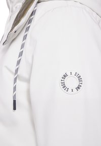 Vit jacka med texturerad tyg, ståkrage och rund logopatch med "STREETONE x STREETONE." Dragsko med rutigt mönster.