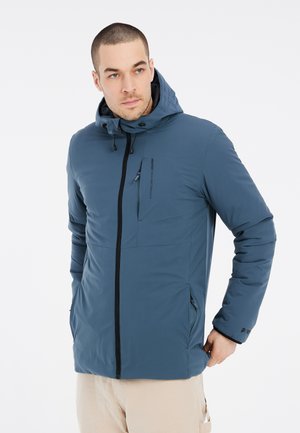 Chaqueta aislante azul con capucha, que presenta un cierre frontal, bolsillo de pecho con cremallera y bolsillos laterales. Textura suave y con un mínimo de marcas.
