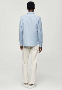 Chemise bleu clair à manches longues avec col classique, coupe ajustée et poignets boutonnés, portée avec un pantalon crème et des chaussures blanches.