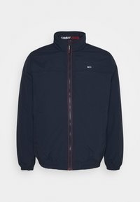 Granatowa kurtka zapinana na zamek z wysokim kołnierzem, elastycznymi mankietami i dołem. Posiada logo na piersi oraz napis "TOMMY JEANS" wydrukowany wewnątrz kołnierza.