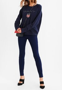 Granatowa bluza z kapturem z logo GANT i herbem, zestawiona z ciemnymi obcisłymi jeansami i czarnymi sandałami na obcasie, z małą torebką na ramię.