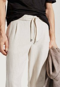 Pantaloni di lino color beige chiaro con vita regolabile tramite coulisse, dotati di pieghe laterali. Nella mano opposta si tiene un giubbotto marrone a trama.