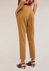 Hnědé tailored kalhoty s hladkou texturou, se bočními kapsami a elegantním střihem, v kombinaci s tmavými sandály na podpatku.