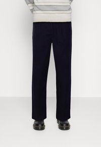 Mørkeblå lige ben bukser med elastisk talje, glat tekstur, og sorte sko med kraftige såler. Stribet sweater synlig ovenfor.