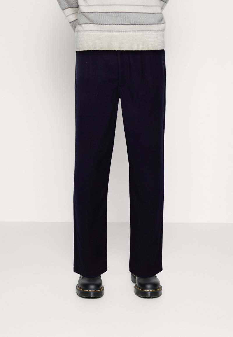 Mørkeblå lige ben bukser med elastisk talje, glat tekstur, og sorte sko med kraftige såler. Stribet sweater synlig ovenfor.