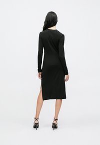 Langermet svart kjole med smal passform, knelang, med sidesplitter. Kombinert med svarte høye hæler. Glatt tekstur.
