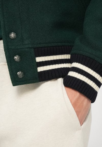 Mână în buzunarul pantalonilor bej deschis, îmbrăcat cu o jachetă verde închis, cu manșete ribbate în dungi negre și albe și trei nasturi metalici.