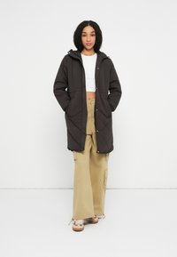 Kvinde iført en lang sort quiltet frakke over en hvid cropped top, beige vide cargo bukser og beige sneakers, stående med hænderne i lommerne.