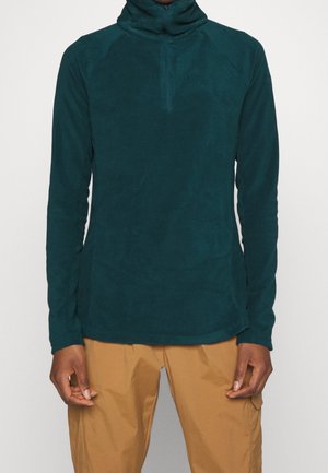 Osoba ubrana w turkusową bluzę polarową z długim rękawem i suwakiem do ćwiartki oraz beżowe spodnie, stojąca na jednolitym tle.