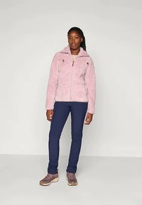 Chlpatá ružová bunda na zips s kapucňou, akcenty so sťahovacou šnúrkou, dve predné vrecká, v kombinácii s námornícky modrými slim-fit nohavicami a hnedými turistickými topánkami.