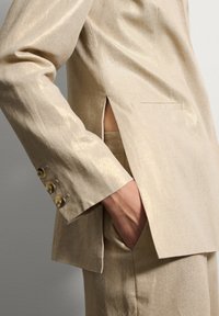 Traje beige a medida con una textura brillante, que cuenta con una abertura lateral y cuatro botones en tono dorado en la manga. Bolsillo visible en el costado.