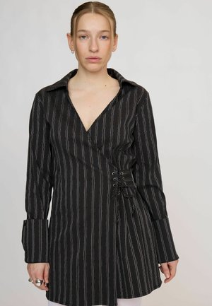 Femme aux cheveux blonds porte une robe portefeuille noire à fines rayures avec un laçage type corset sur le côté et de larges manches, debout devant un fond uni.
