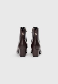 Botas de cuero marrón hasta el tobillo con un tacón medio en bloque, que cuentan con una cremallera lateral y un logo acentuado en dorado en la parte trasera. Textura suave y diseño elegante.
