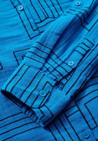 Chemise bleue à manches longues avec motifs de lignes noires verticales et carrées et boutons bleus assortis aux poignets et sur le devant.