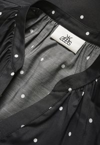 Sort bluse med hvide prikker, der har et blødt, tekstureret indre og et mærkat. Formen inkluderer en plisseret halsdesign.