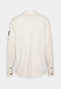 Jasnobeżowa koszula z długim rękawem, wykonana z bawełny, klasyczny kołnierz, mankiety na guziki oraz czarna materiałowa naszywka na lewym ramieniu.