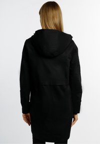 Personne aux cheveux châtains clairs et raides, portant un long manteau à capuche noir, debout de dos sur un fond blanc.