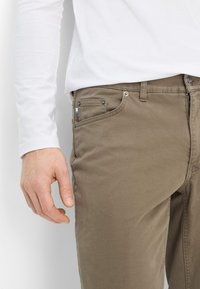 Braune Baumwollhose mit Reißverschluss, Vorderseite mit Taschen und kontrastierenden Nähten. Getragen mit einem weißen Langarmhemd, das eine lockere Passform zeigt.