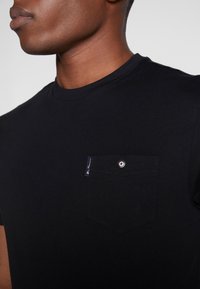 Camiseta de algodón negra con cuello redondo, que presenta un bolsillo en el pecho con un botón en contraste y una etiqueta de logo. Textura suave.