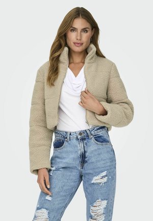 Beige, cropped fleece jack met hoge kraag, ritsluiting en gestructureerd oppervlak, gecombineerd met versleten blauwe jeans en een witte top.