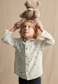 Hellgrünes, knöpfbares Hemd mit kleinen Tier- und Pflanzendrucken; getragen von einem Kind, das einen plüschigen beigen Teddybären über den Kopf hält.