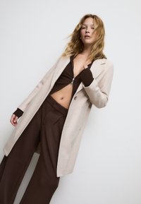 Manteau long beige avec revers crantés porté sur un haut zippé marron et un pantalon marron. Tissu texturé et coupe décontractée, avec une fermeture à un bouton.