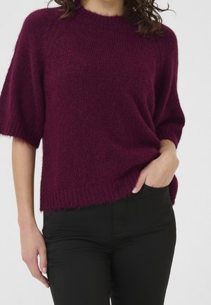 Femme portant un pull violet duveteux à manches courtes et amples, rentré dans un pantalon noir ajusté, une main dans la poche et cheveux bouclés visibles.