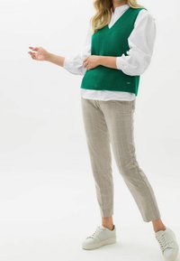 Veste en tricot côtelé vert portée sur une chemise blanche, associée à un pantalon slim à carreaux gris et des baskets blanches. La veste présente un col en V.
