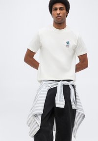 Άνδρας που φοράει λευκό πουκάμισο με κοντά μανίκια και μικρό λογότυπο, μαύρο παντελόνι και μπλε-λευκή ριγέ μπλούζα με μακριά μανίκια δεμένη γύρω από τη μέση.