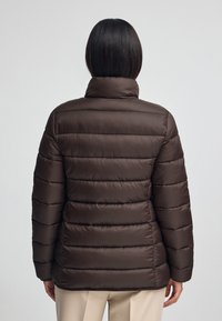 Brun puffjacka med quiltad design, hög krage och mjuk textur. Har sydda horisontella paneler och en figurnära silhuett.