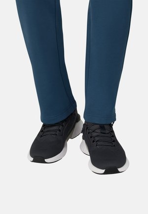 Schwarze Sportsneaker mit strukturiertem Mesh-Obermaterial und weißer Sohle, kombiniert mit dunkelblauen, schmal zulaufenden Jogginghosen mit glatter Oberfläche.