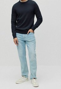 Uomo che indossa un maglione nero a maniche lunghe, jeans azzurri chiari e sneakers bianche, in piedi con una mano in tasca davanti a uno sfondo semplice.