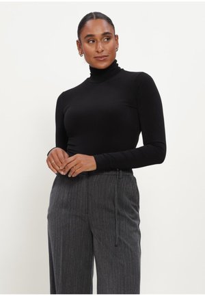 Woman wearing black long-sleeve turtleneck and high-waisted gray pinstripe pants, posing with hands clasped in front on light background.