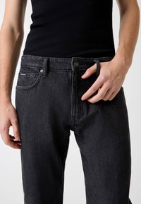 Jean en denim noir coupe droite, avec fermeture par boutons, poches latérales et étiquette de marque visible à l'arrière. Texture douce.