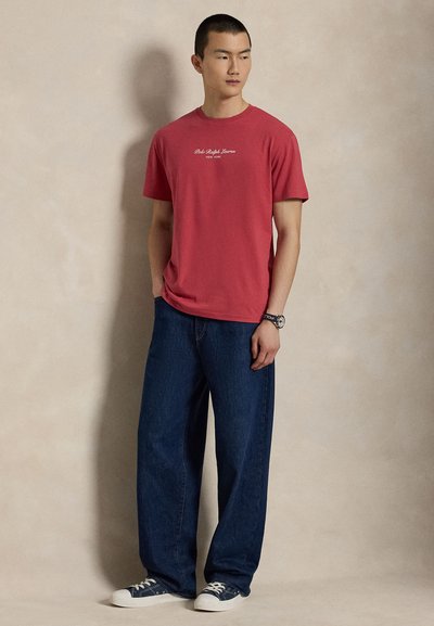 Νέος άνδρας που φοράει κόκκινη μπλούζα Polo Ralph Lauren, φαρδιά σκούρα μπλε τζιν, ναυτικό μπλε αθλητικά παπούτσια και ρολόι χειρός, με φόντο απλό φωτεινό.