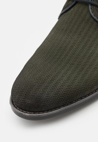 Scarpa in suede verde scuro con una texture a zigzag, punta arrotondata e suola scura a contrasto. Presenta una allacciatura minimalista per la chiusura.